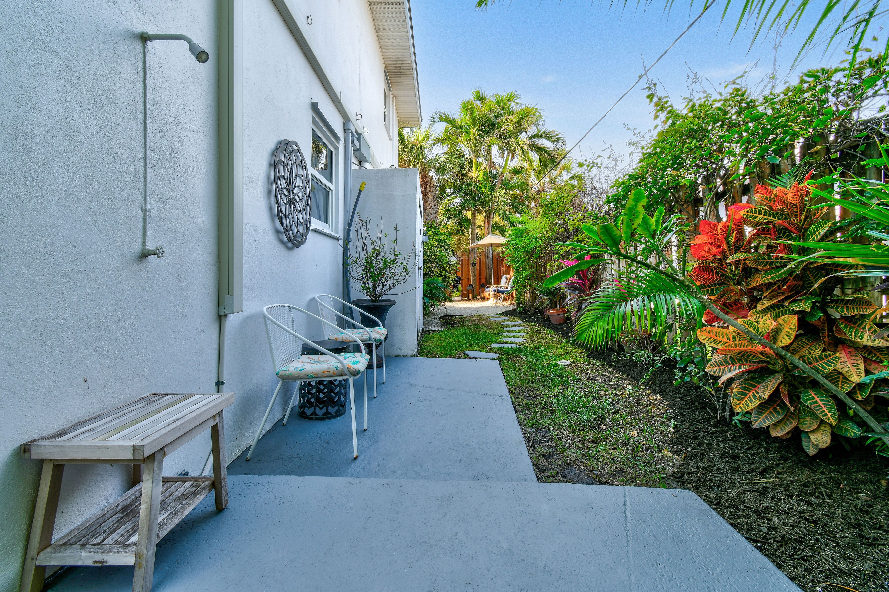 A small outdoor seating area with a bench and chairs, a concrete pathway, and lush tropical plants.