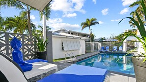 Zona de piscina con sillones de descanso, una palmera y un edificio al fondo.