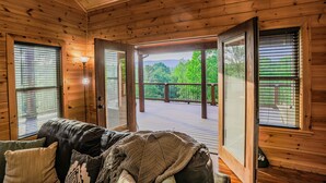 Ein Holzhütten-Innenraum mit einer Couch, einer Decke und Blick auf eine Terrasse und Grünflächen.