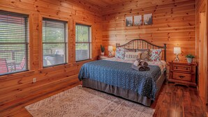 Ein Schlafzimmer einer Holzhütte mit einem Bett, zwei Nachttischlampen, einem Nachttisch und einem Fenster mit Jalousien.