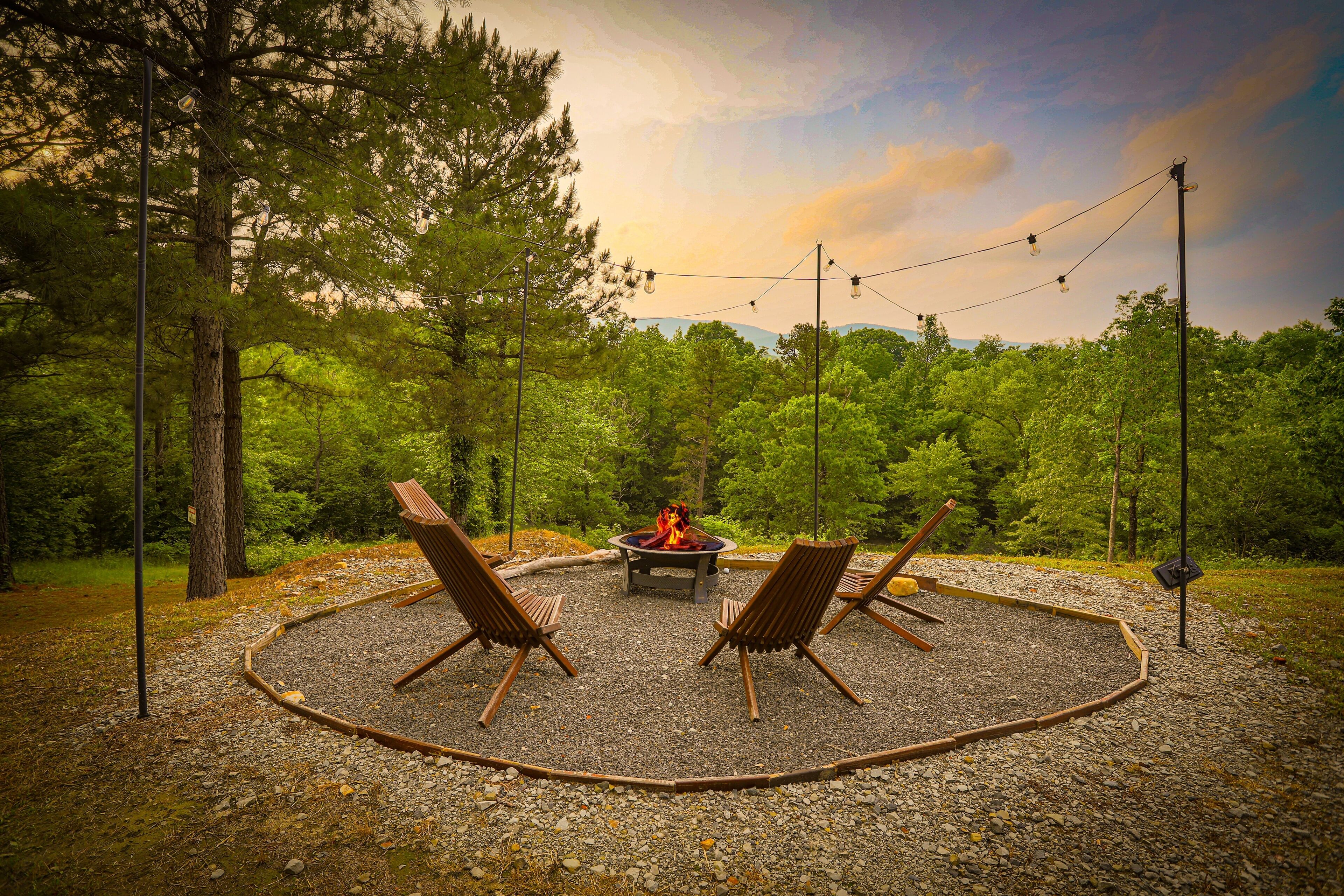 Un espace circulaire aménagé avec un foyer, entouré d’un chemin de gravier et de chaises en bois, situé dans une zone boisée et illuminé par des guirlandes lumineuses suspendues au-dessus.