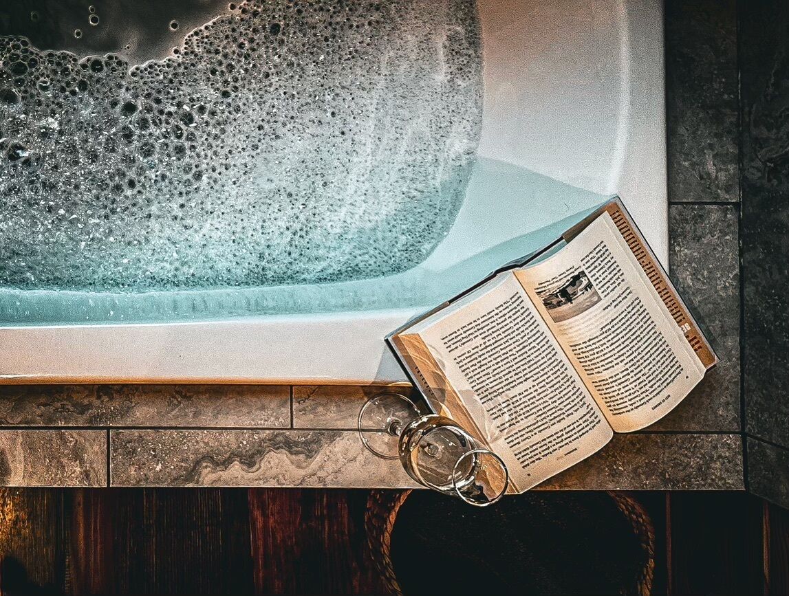 Una bañera con burbujas, un libro y dos copas de vino sobre una superficie de madera.