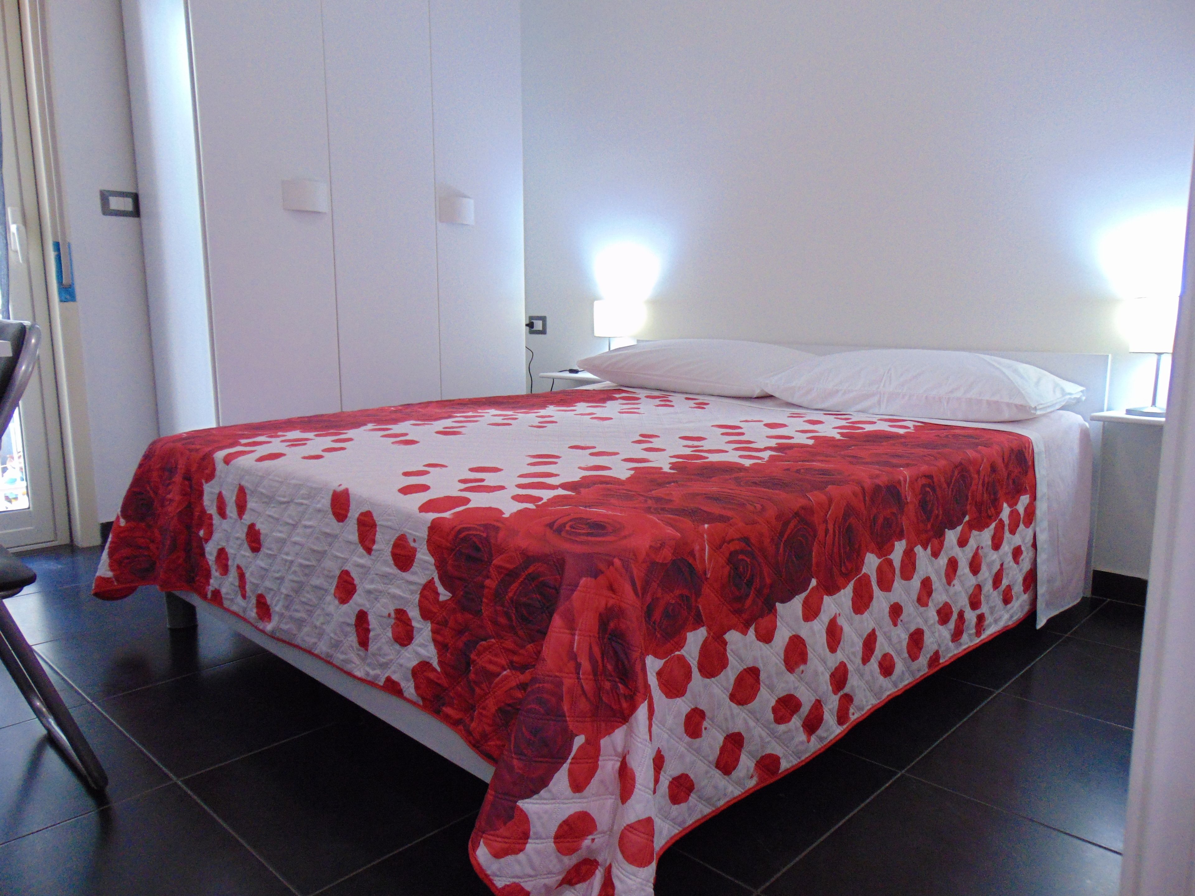 A neatly made bed with a red and white floral bedspread, a white pillow, and a white headboard.