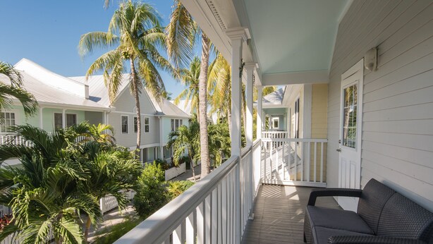 A covered porch with white railings, a wicker sofa, and a view of a tropical landscape with palm trees.