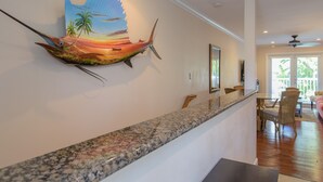 A kitchen with a granite countertop, a dining area with wicker chairs, and a wall-mounted artwork of a tropical scene.