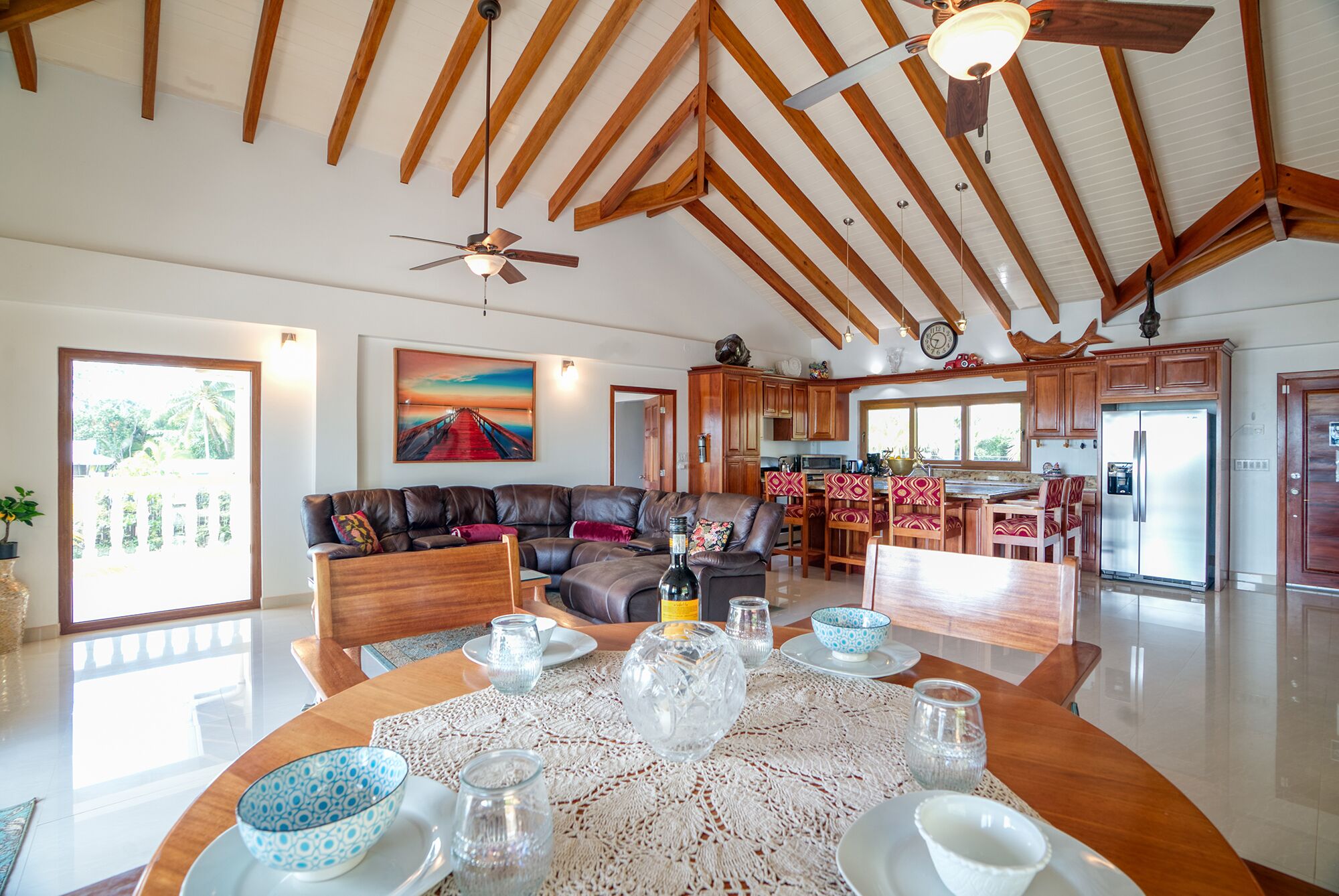A spacious living area with a high ceiling and wooden beams, a dining table set with glassware, and a kitchen with wooden cabinets and a refrigerator.