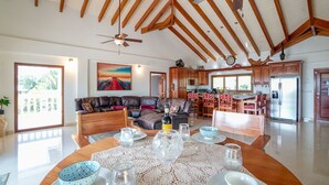 A spacious living area with a high ceiling and wooden beams, a dining table set with glassware, and a kitchen with wooden cabinets and a refrigerator. - Beachfront Villa • Private Pool • Fully Fenced • Walk to Restaurants (Placencia)