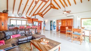 A spacious living room with a leather sofa, a glass coffee table, and wooden chairs. A kitchen with wooden cabinets and a dining area with wooden chairs are visible in the background. - Beachfront Villa • Private Pool • Fully Fenced • Walk to Restaurants (Placencia)
