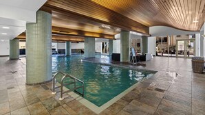 An indoor swimming pool with tiled walls and wooden ceiling beams, surrounded by stone flooring and a glass door leading outside.
