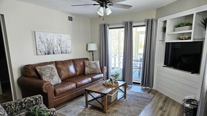 Ein Wohnzimmer mit braunem Ledersofa, gemusterten Sesseln, einem Holztisch und einem Flachbildfernseher, der an einer weißen Wand befestigt ist.