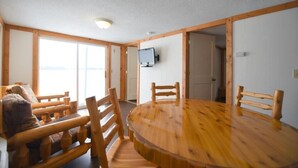 Een kamer met een ronde houten tafel, houten stoelen, een aan de muur bevestigde televisie en een deur naar een andere kamer.