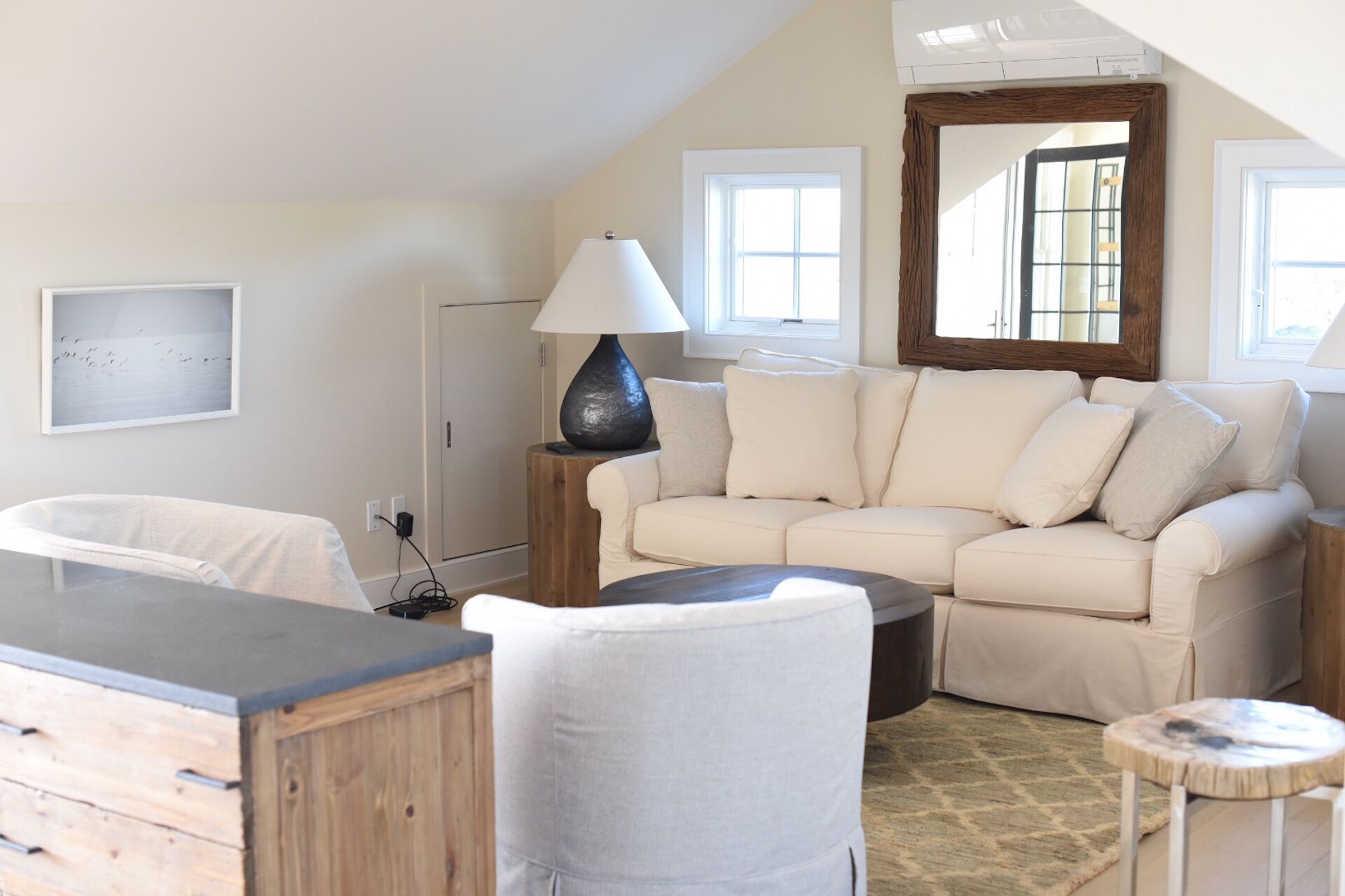 A cozy living room with a beige sofa, a wooden coffee table, and a lamp. There is a white chair and a small round table with a rug.