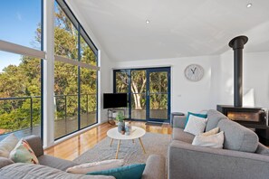 A modern living room with a large TV, a fireplace, and a view of trees through large windows.