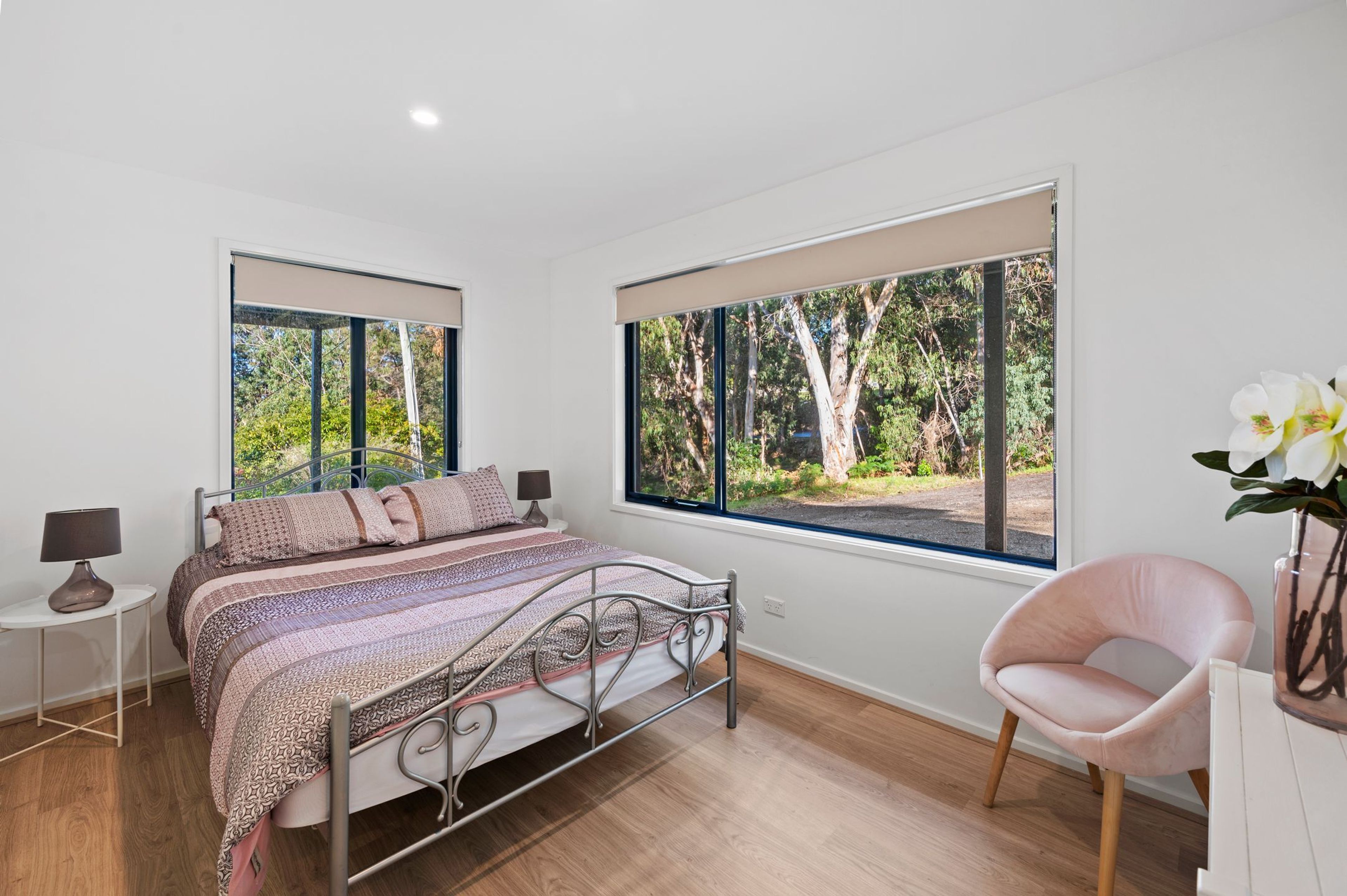 Une chambre à coucher comprenant un lit, des tables de chevet, une chaise et de grandes fenêtres donnant sur la forêt.