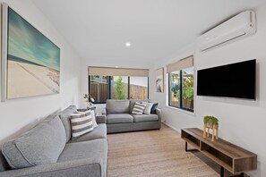 Un salon moderne avec un canapé gris, un téléviseur à écran plat fixé au mur et une grande fenêtre offrant une vue sur l’extérieur.