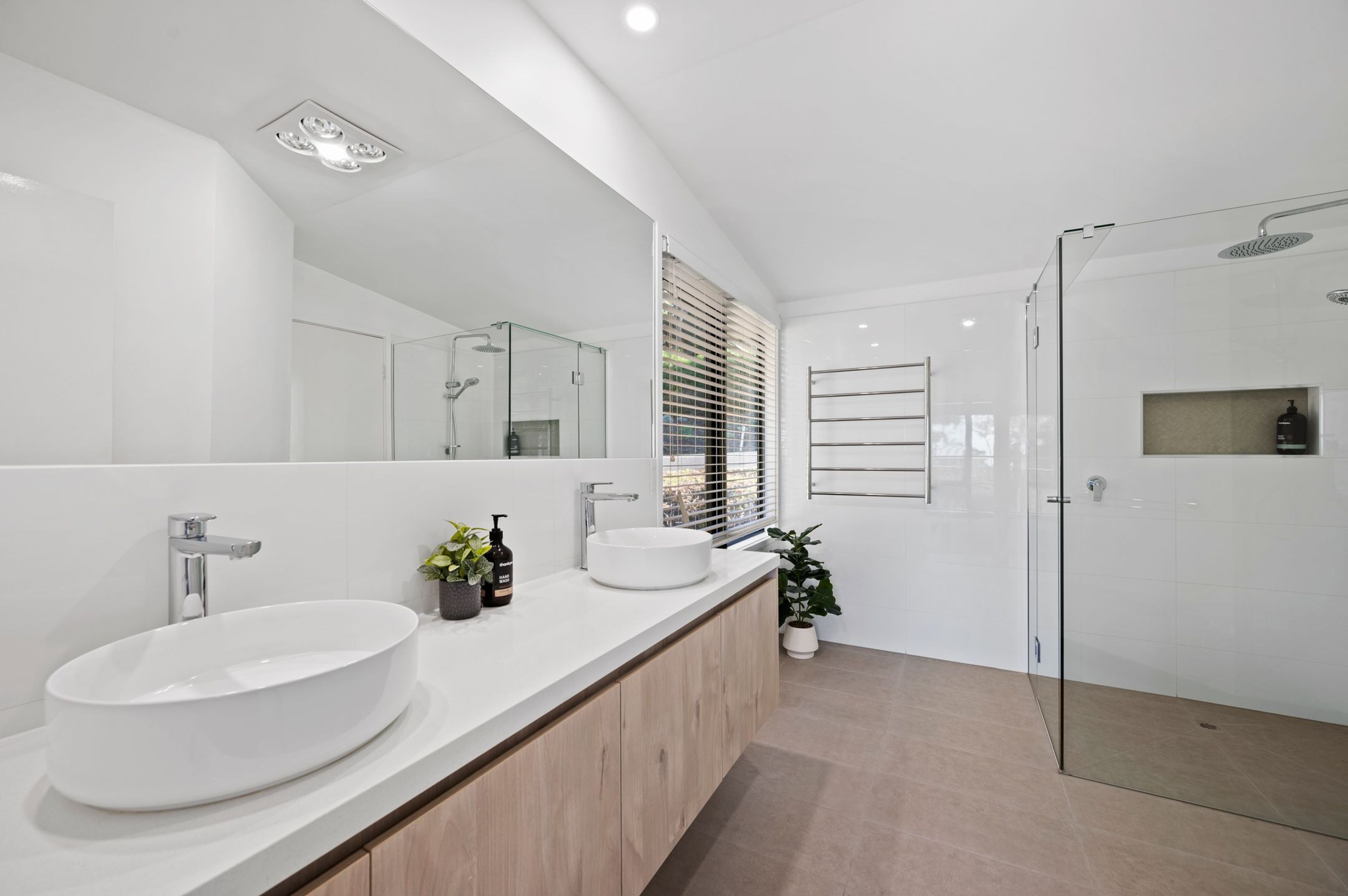 A modern bathroom with a large mirror, two sinks, a shower, and a potted plant.