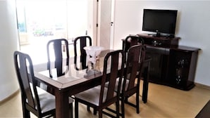 A dining area with a wooden table and chairs, a white vase, and a television on a cabinet. - Casa Maceio Shopping (Maceió)
