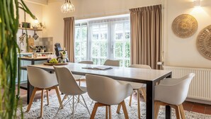 A modern dining area with a large table, white chairs, and a view of greenery outside.