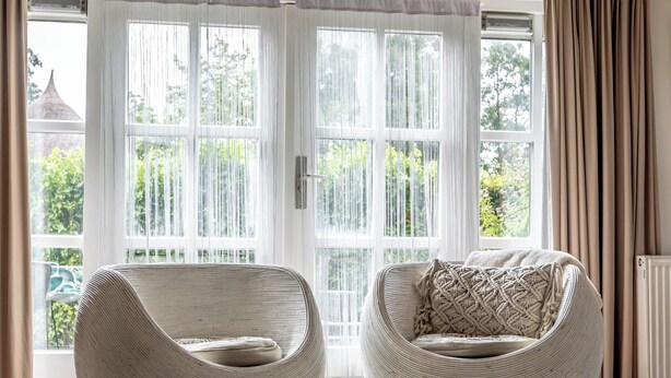 A room with two wicker chairs, a small table, and a large window with curtains.