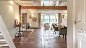 A spacious living area with a high ceiling, exposed wooden beams, a dining table with chairs, a sofa, and a large window with curtains.