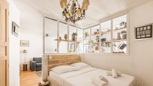 Ein Schlafzimmer mit einem Holzkopfteil, einem großen Bett mit weißer Bettwäsche, einer Chandelier und Blick auf Küchenregale durch ein Fenster.