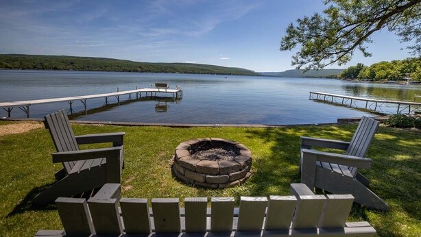 A lakeside deck with Adirondack chairs, a fire pit, and a dock extending into the water. - The North Star of Honeoye: Family Friendly, Great View of Honeoye Lake! (Honeoye)