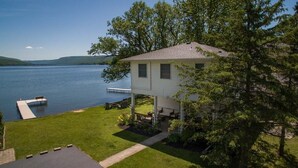 A two-story house with a dock extending into a lake, surrounded by trees and a well-maintained lawn. - The North Star of Honeoye: Family Friendly, Great View of Honeoye Lake! (Honeoye)