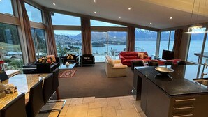 A modern kitchen with a large island, a dining area with a red sofa, and a view of a lake and mountains through large windows. - 7Manata - Amazing 270deg views! Golf & Ski 10bedrm/8bathrm + 4x seven-seat 4WD's (Fernhill)