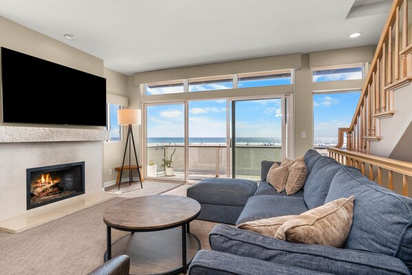 A modern living room with a fireplace, a flat-screen TV, a sofa, and a balcony with a view of the beach.