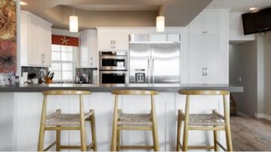 A modern kitchen with white cabinets, stainless steel appliances, and three wooden bar stools. - Seaside Home - Full home rental - Beachfront, Full Beach Front Home - 2002 W. Oceanfront (Newport Beach)