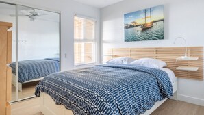 Un dormitorio con una cama grande, cabecera de madera, una ventana con persianas, un ventilador de techo y un cuadro de un barco en la pared.