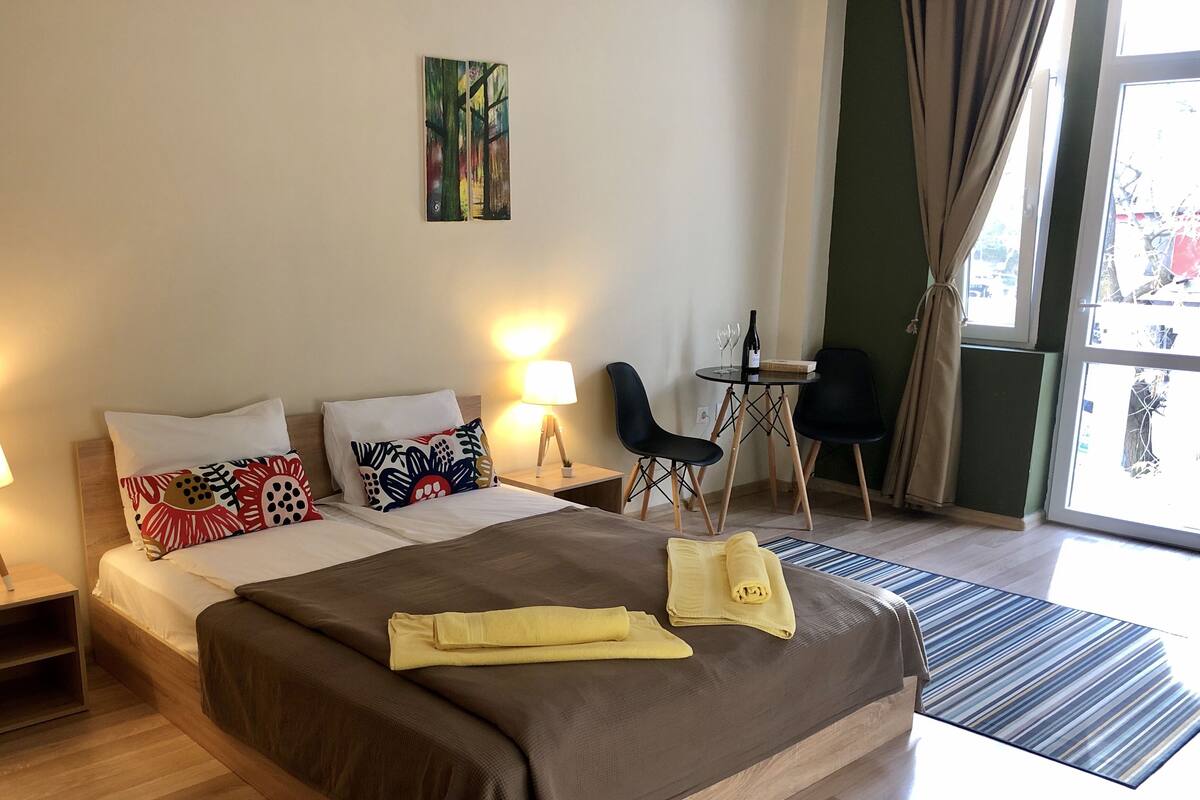 A hotel room with a large bed, two bedside tables with lamps, a small table with a bottle and glasses, a chair, and a view through the window.