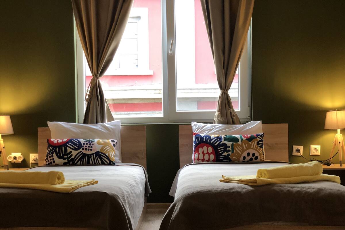 A hotel room with two beds, each with a patterned pillow and a rolled towel. There are bedside lamps and a window with curtains.
