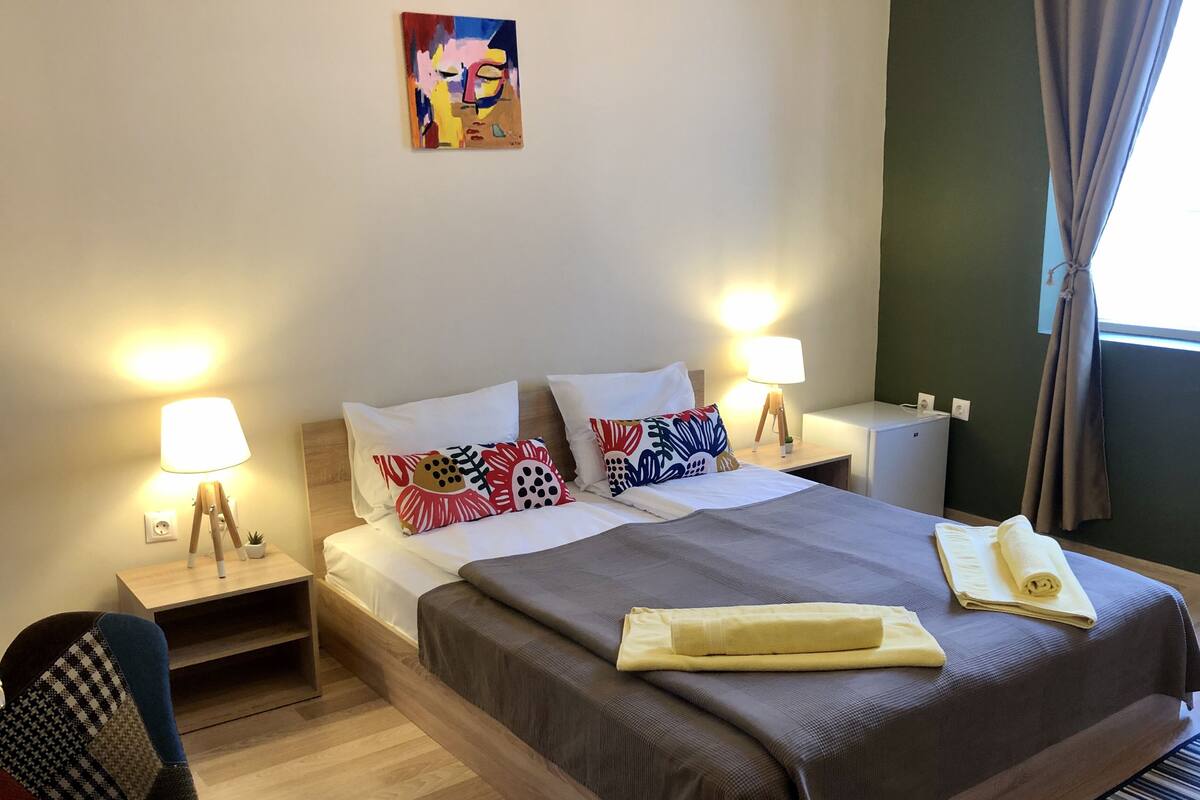 A hotel room with a large bed, two bedside tables with lamps, a small desk, a potted plant, and a painting on the wall.
