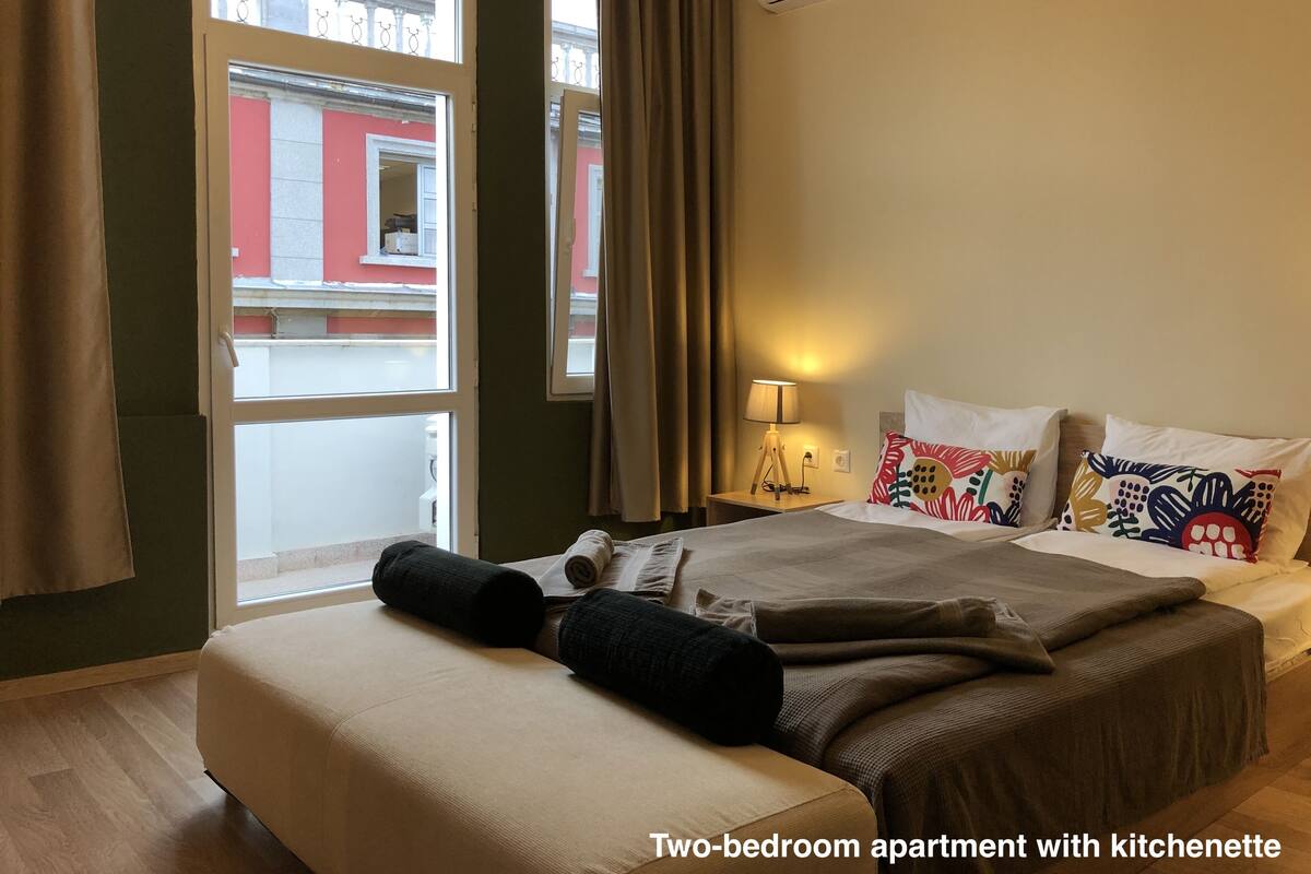 A two-bedroom apartment with a kitchenette, large windows, and a view of the street.