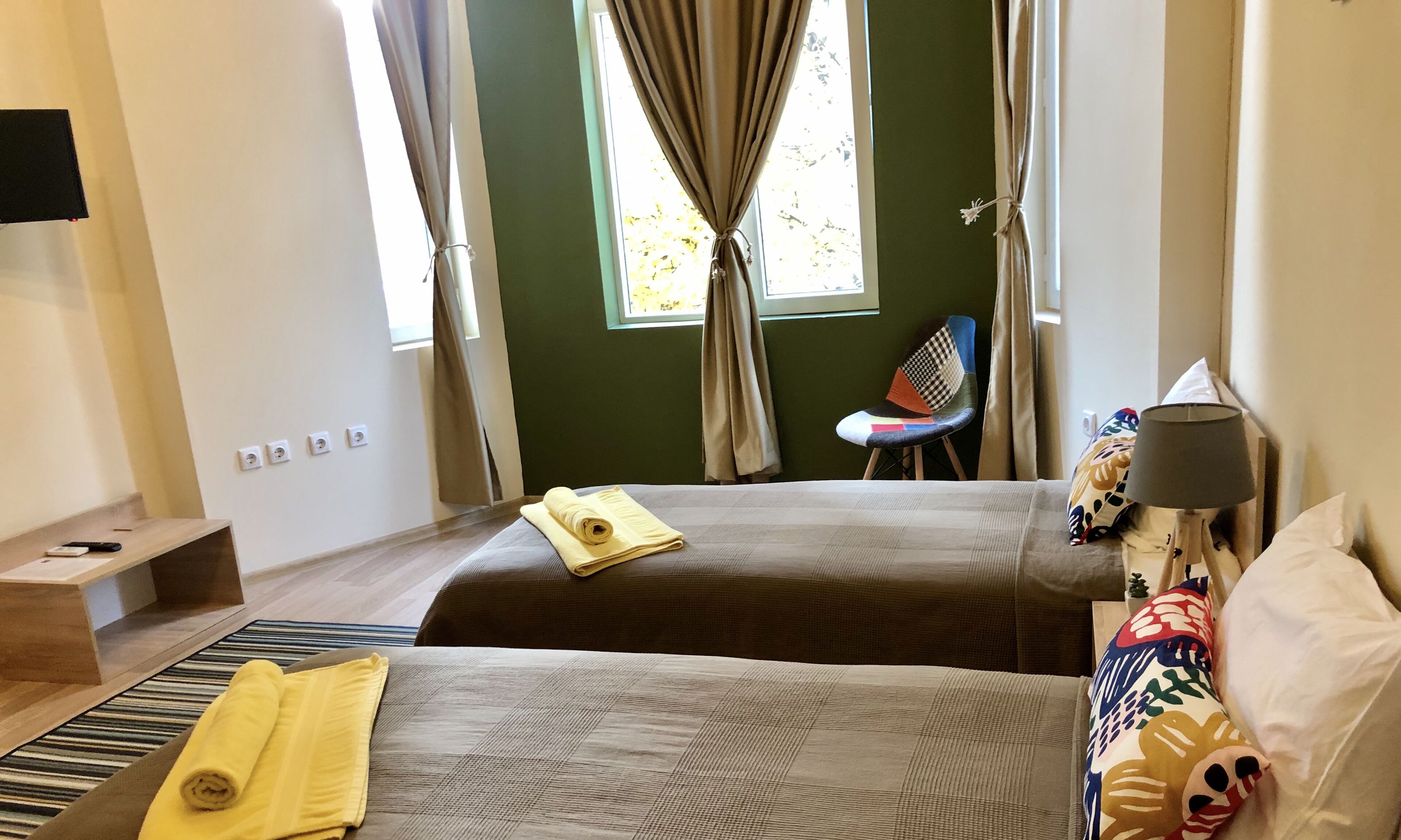 A hotel room with two beds, a chair, a lamp, and a TV on the wall.