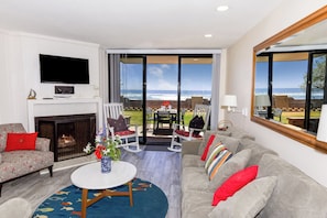 A living room with a fireplace, a flat-screen TV, and a view of the ocean.