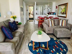 A living room with a grey sectional sofa, a round coffee table, and a colorful rug. There is a dining area with red chairs and a kitchen in the background.