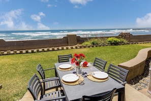 An outdoor dining area with a view of the ocean, a warning sign about unstable rocks, and a table set for two with a vase of red flowers.