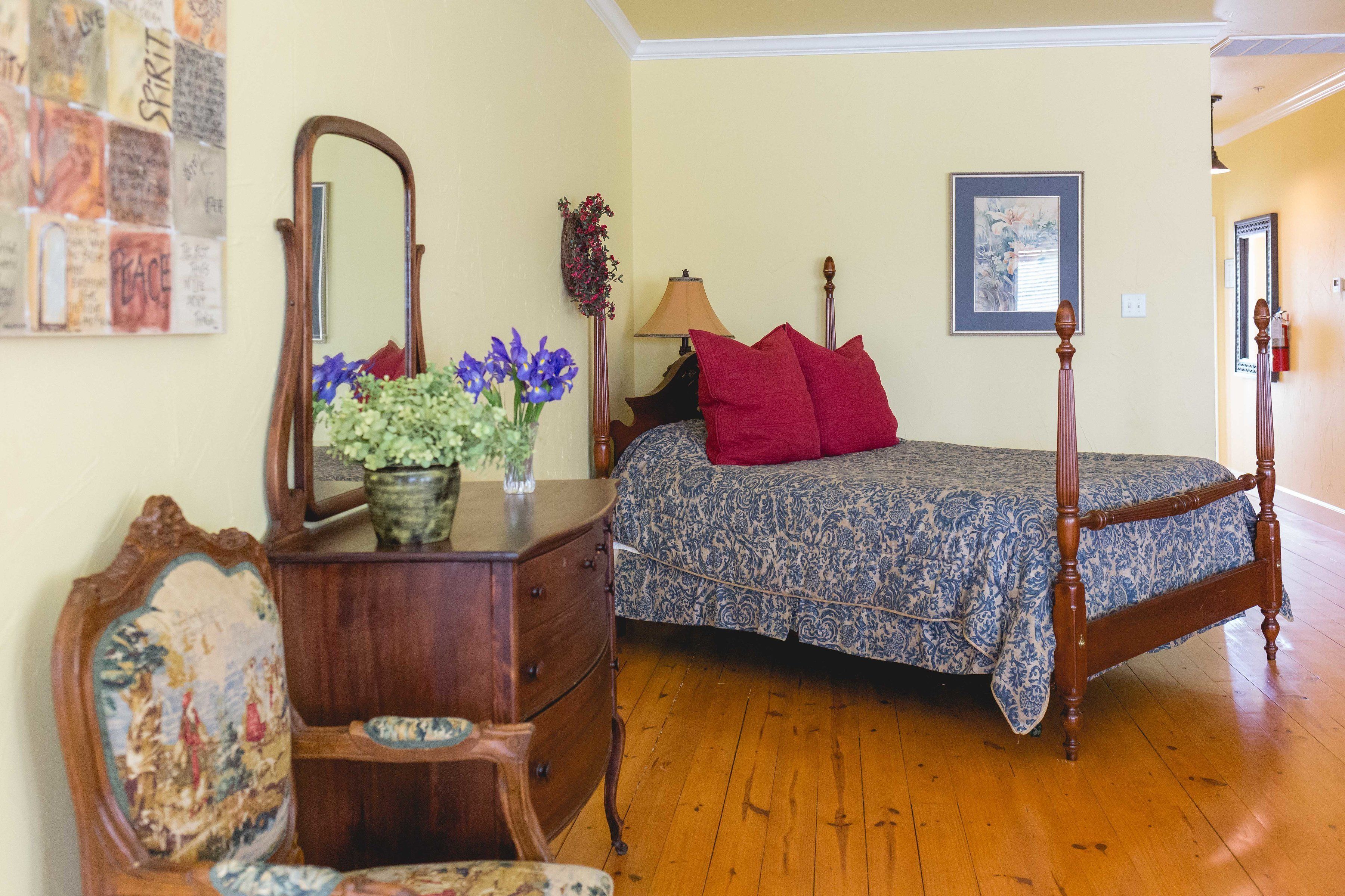 A bedroom with a wooden four-poster bed, a dresser, a chair, a mirror, and a wall with a framed picture and a bulletin board.