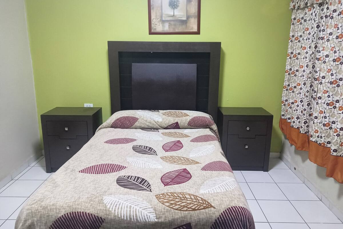 A bedroom with a bed, two nightstands, a headboard, a framed picture on the wall, and a patterned curtain.