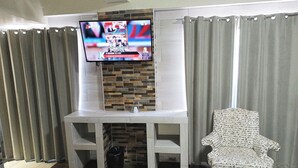 A living room with a TV mounted on a brick wall, a white console table, a patterned armchair, and a black trash bin. - Hotel Baeza (Delicias)