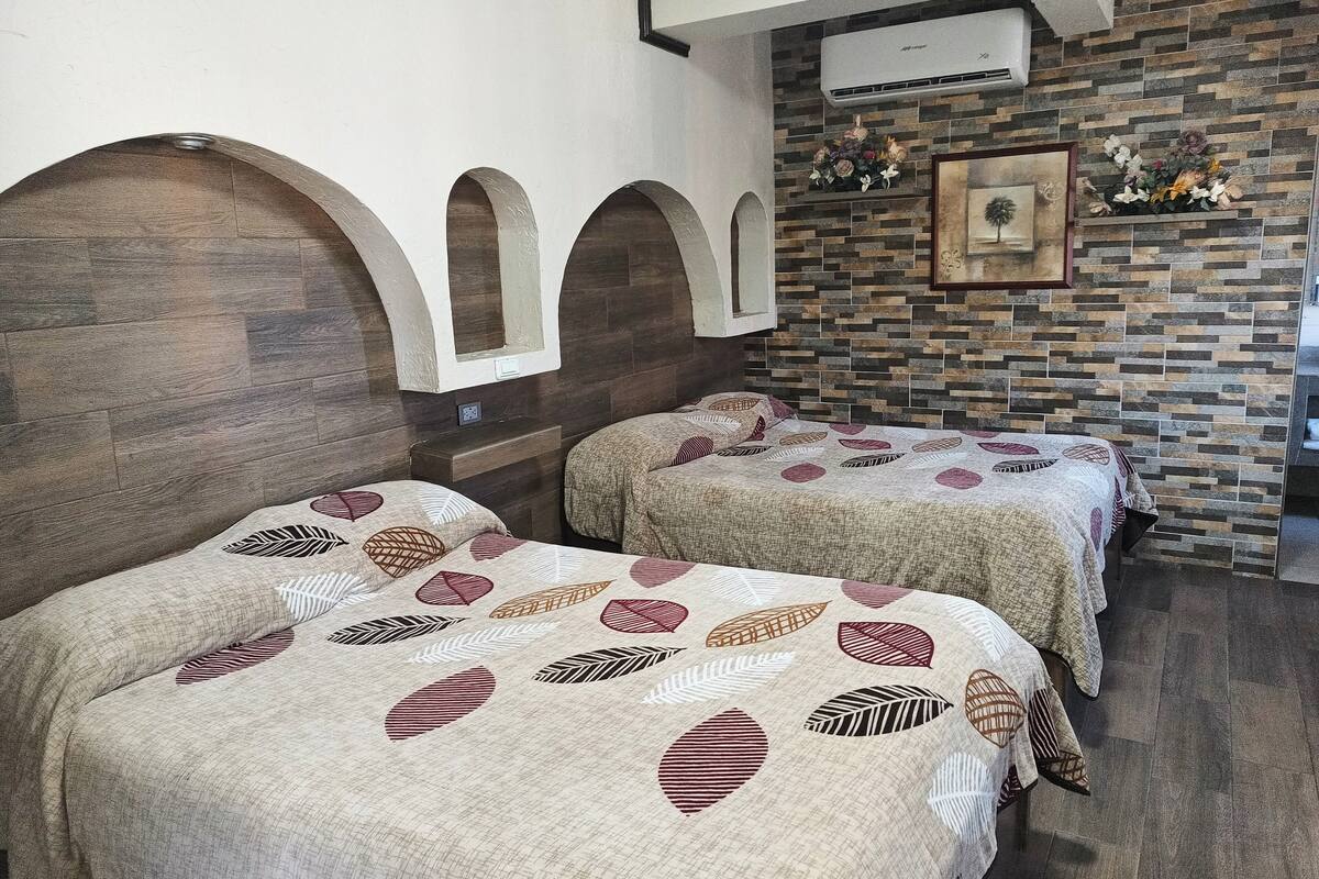 A room with two beds, each with patterned bedding. There is a stone wall with a floral arrangement and a framed picture. An air conditioning unit is mounted on the wall.