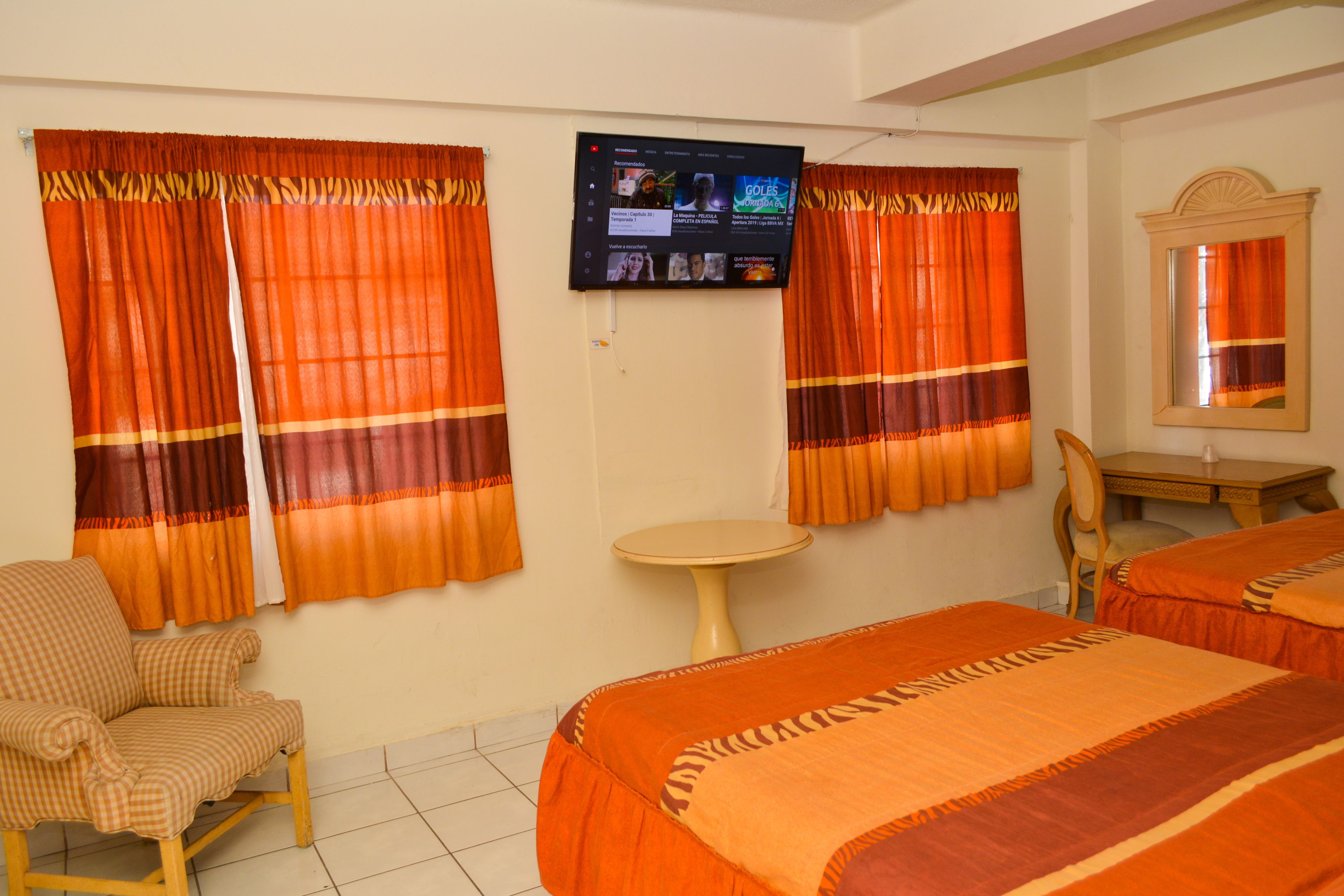 A hotel room with two beds, a television mounted on the wall, a small round table, a chair, and curtains with a striped pattern.