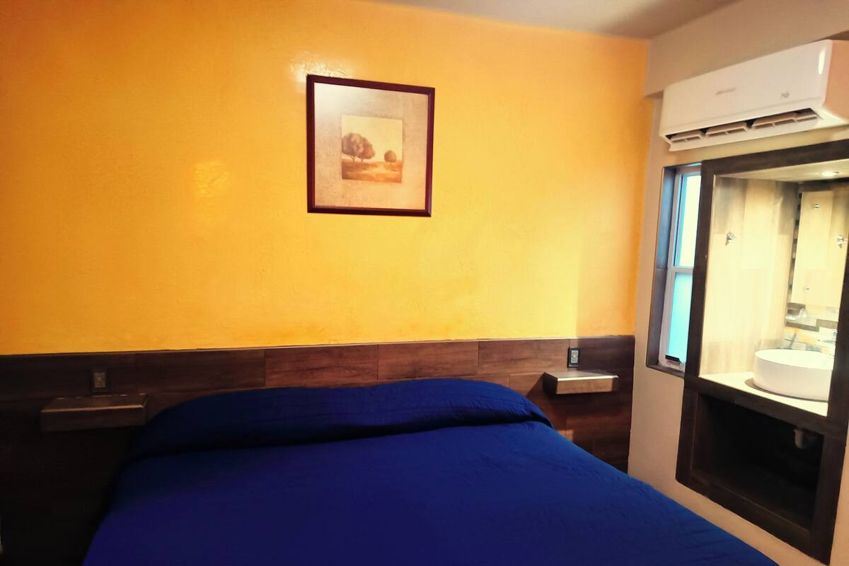 A hotel room with a bed, a framed picture on the wall, a nightstand, and a window with a view of a bathroom.