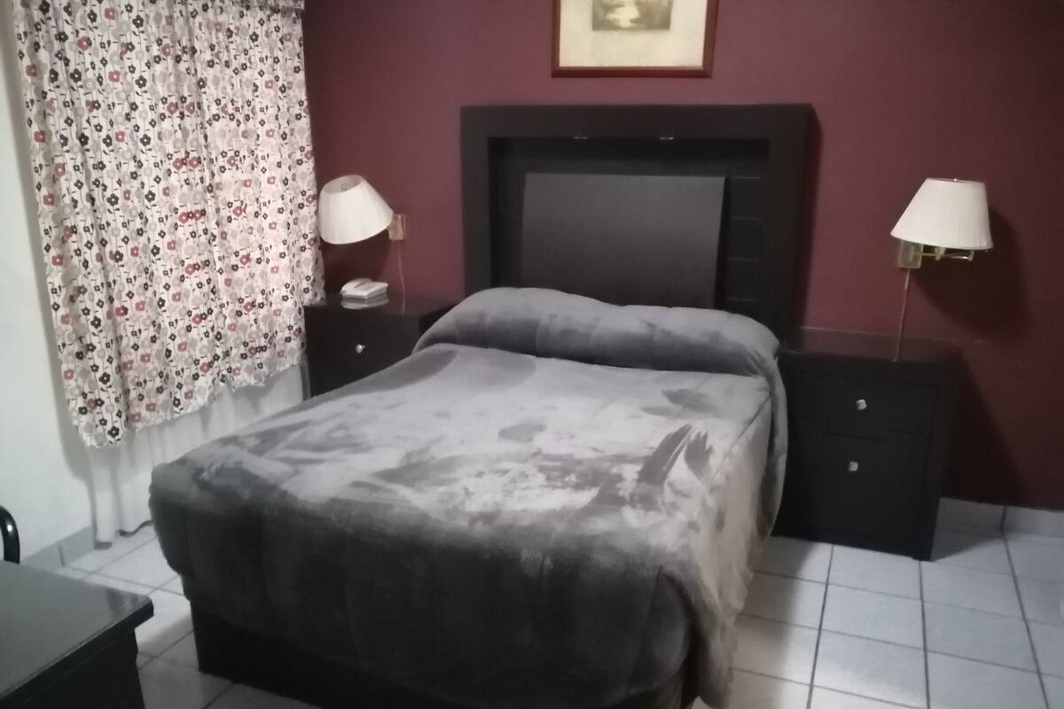 A bedroom with a bed, two bedside tables, a nightstand, a lamp, a curtain, and a framed picture on the wall.