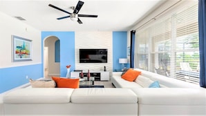 A modern living room with a white sofa, orange cushions, a flat-screen TV, and a ceiling fan.