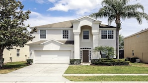 A two-story house with a white garage door, a central entrance with a brown door, and a small front yard with a palm tree and shrubs.