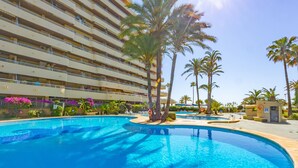 A large outdoor swimming pool surrounded by palm trees and a multi-story hotel building. - Apartment Turmalina - PlusHolidays (Calpe)