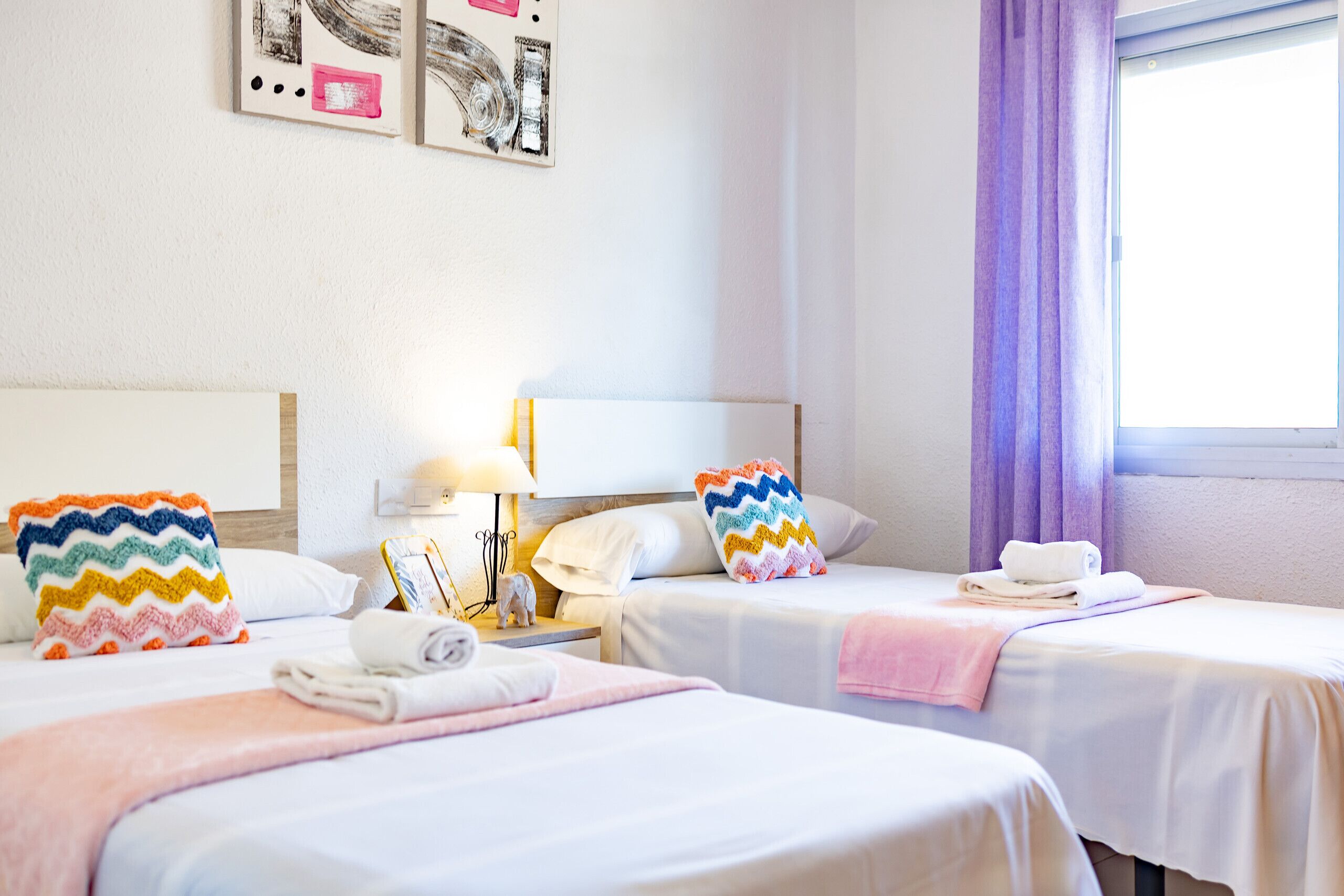 Ein Hotelzimmer mit zwei Betten, jedes mit einem farbenfrohen Kissen, weißer Bettwäsche und einem kleinen Nachttisch mit Lampe und Dekoration.